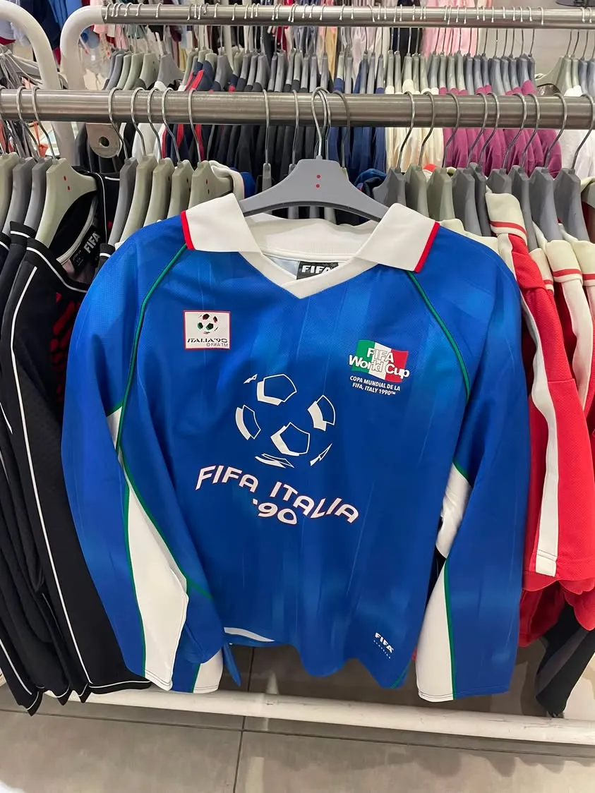 Cotton On 店里衣架上一件蓝色 FIFA Italia '90 复古长袖球衣,胸前有 Italia '90 标志和 FIFA World Cup 意大利国旗徽章