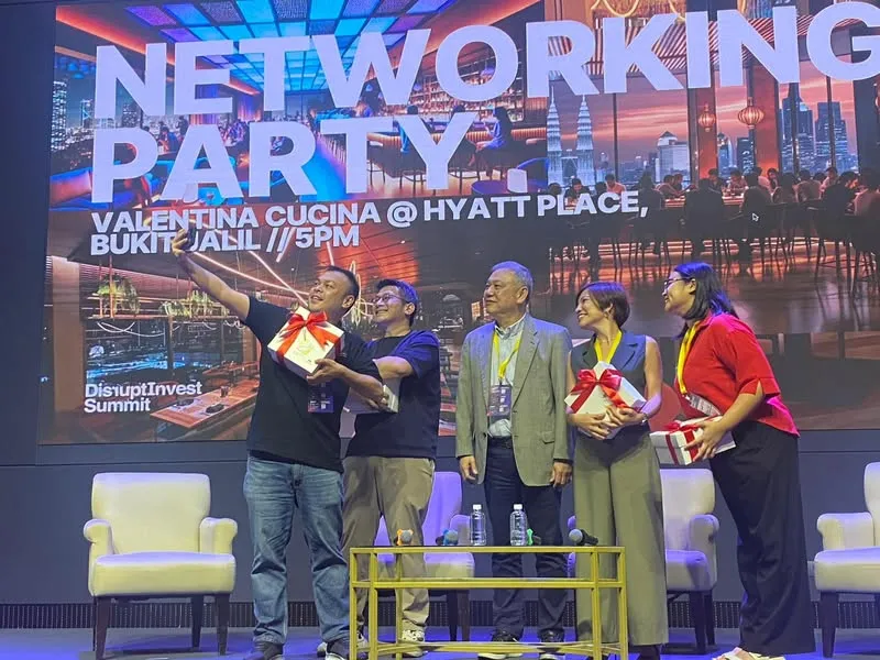 Speakers and the host on the DisruptInvest Summit stage taking a selfie while holding gifts, with a big screen behind them reading Networking Party, Valentina Cucina @ Hyatt Place, Bukit Jalil