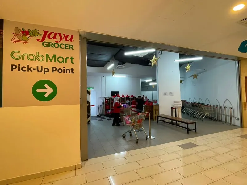 商场里 Jaya Grocer GrabMart Pick-Up Point 的入口,里面员工在推着购物车整理订单,旁边一排红色袋子和购物车