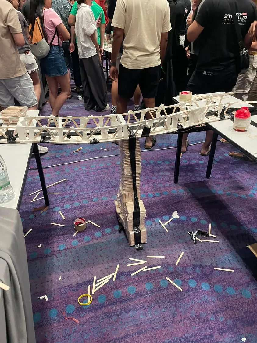 A bridge made of popsicle sticks and tape at NEXEA Entrepreneur Camp, spanning two tables and propped up in the middle by a stack of sticks wrapped in black tape, with loose sticks scattered on the carpet