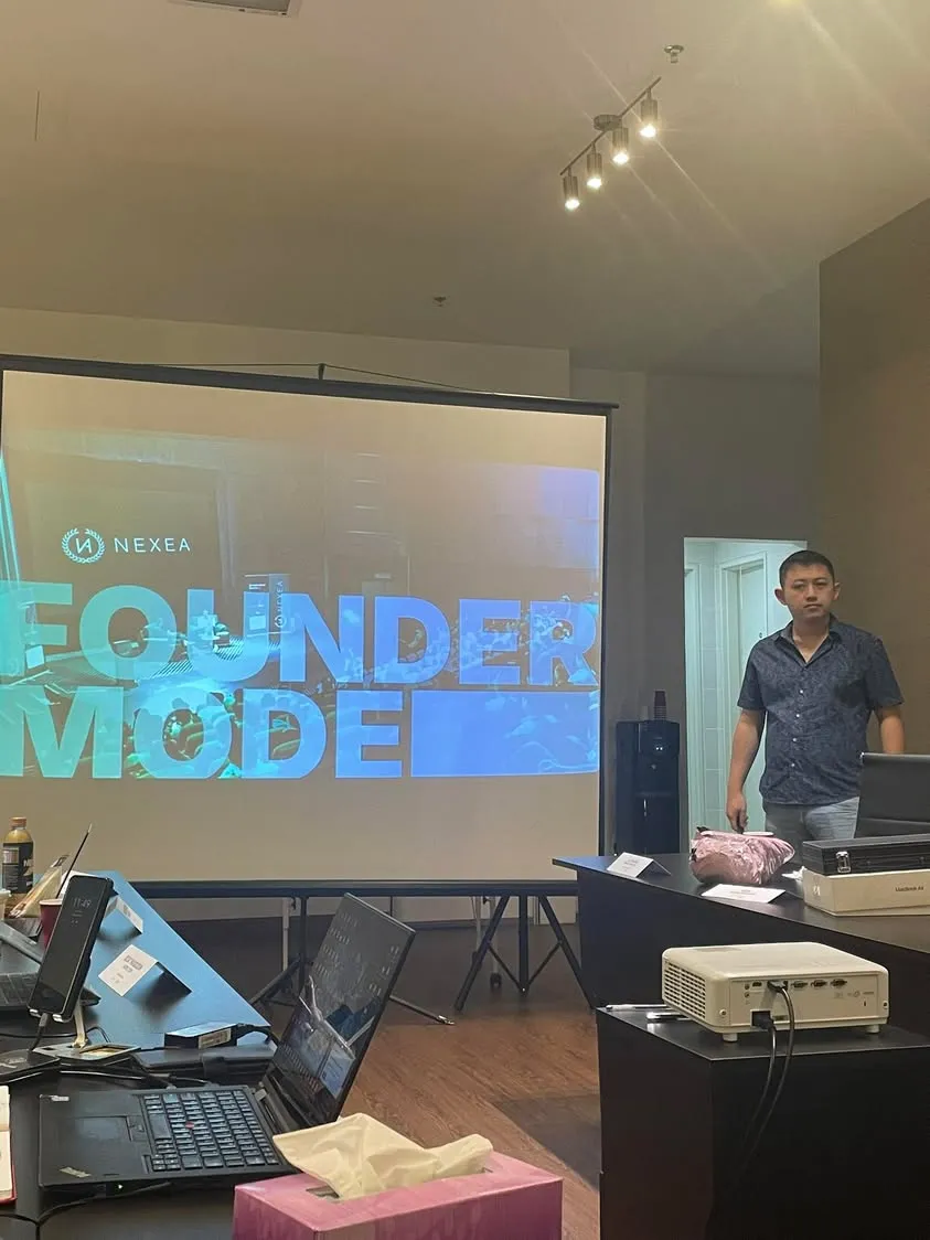 A NEXEA Founder Mode session, with "NEXEA FOUNDER MODE" on the projector screen and the speaker standing at the front of a meeting room, laptops and a projector on the tables