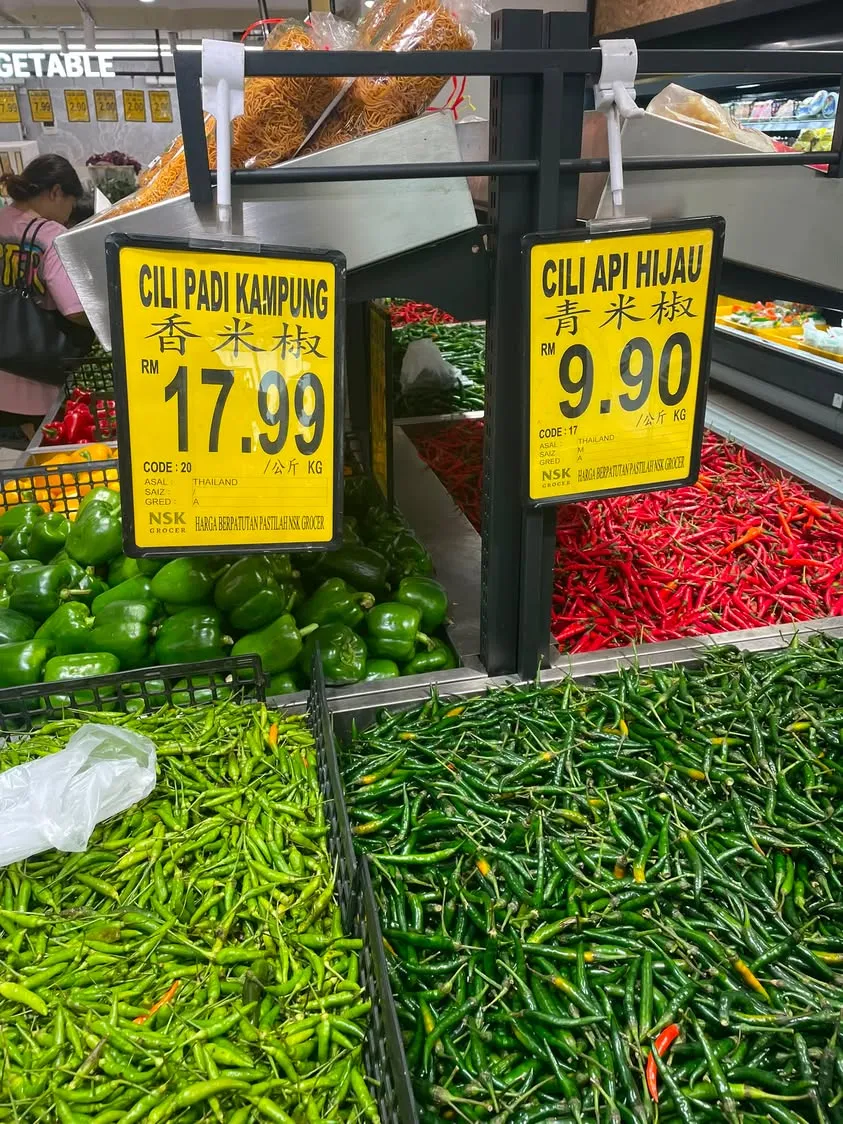 NSK 菜市场的两块黄色价格牌:Cili Padi Kampung 香米椒 RM17.99/kg 和 Cili Api Hijau 青米椒 RM9.90/kg,产地都写 Thailand,下面是成堆的青辣椒和红辣椒