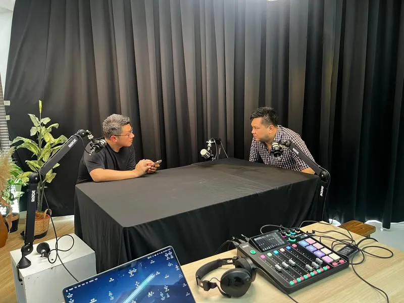 podcast 录音室里,两个人隔着铺黑布的桌子对着 RØDE 麦克风聊天,前景是混音台、耳机和笔记本电脑