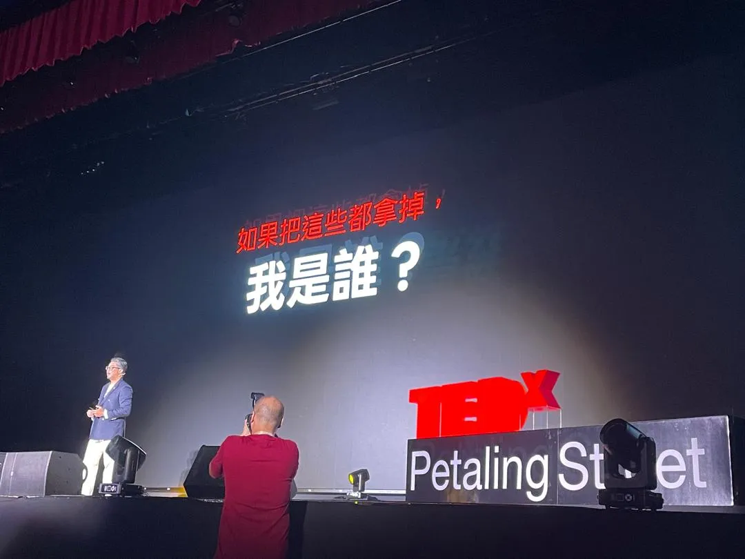 TEDxPetalingStreet 台上,讲者站在舞台左边,背后大屏幕写着「如果把这些都拿掉,我是谁?」,台前有摄影师在拍照