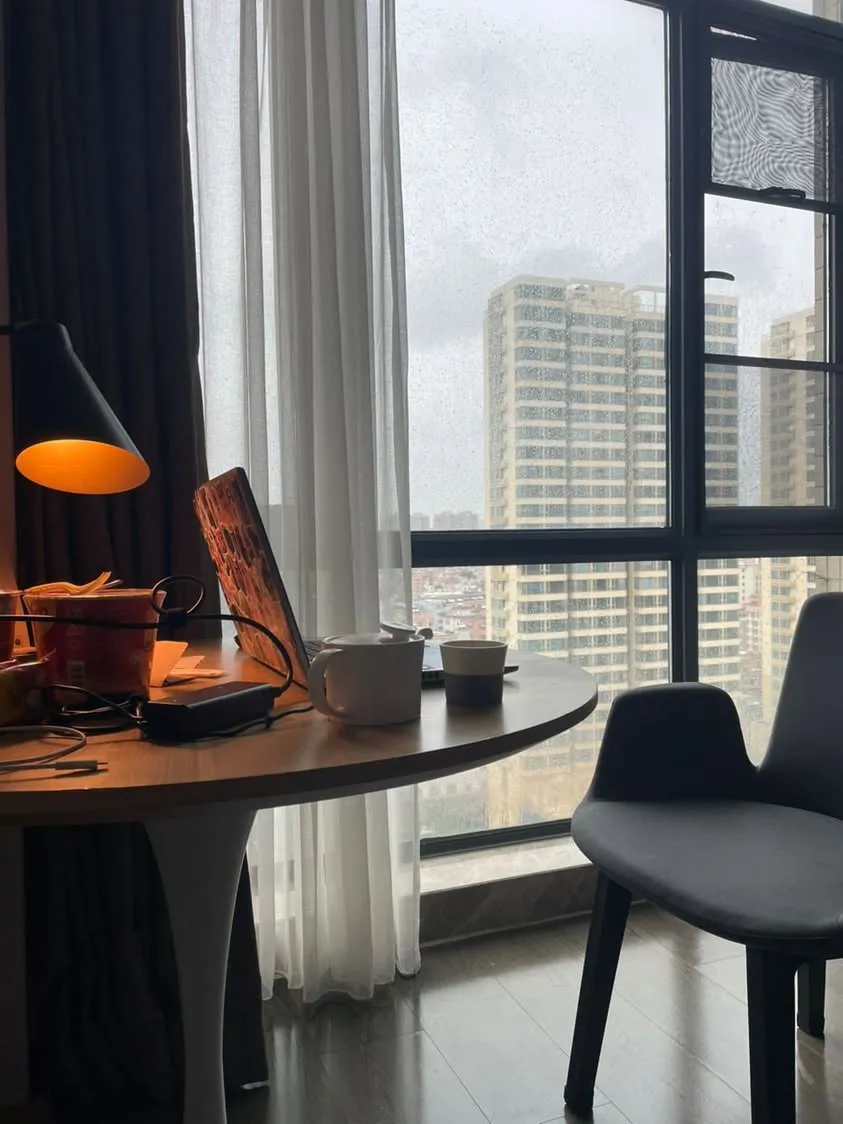 Stuck in a hotel room during the typhoon: rain on the window, a desk with a lamp, a laptop, a teapot and a cup of instant noodles, and high-rise blocks outside
