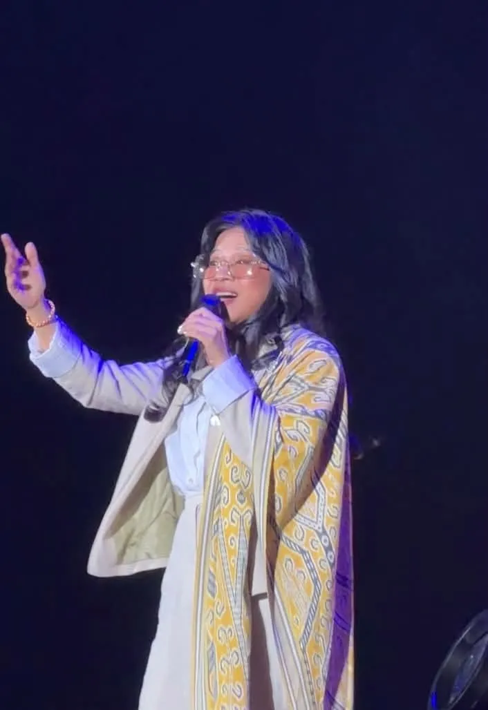 Zee Avi on stage at TEDxPetalingStreet, singing into a microphone in a coat with a yellow traditional pattern, against a dark blue background